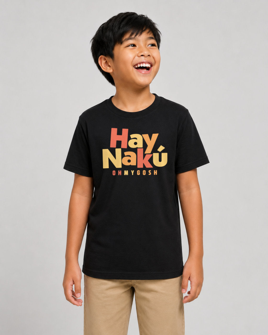A young boy smiling and looking to the side, wearing a black t-shirt with a colorful graphic that reads 'Hay Nakú' and 'OH MY GOSH' in orange and yellow, paired with khaki pants, against a solid gray background.