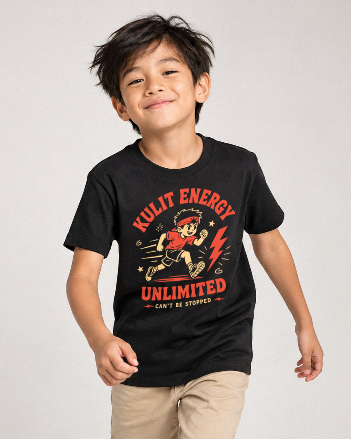 A young boy wearing a black graphic t-shirt with a red and yellow cartoon character and the text 'KULIT ENERGY UNLIMITED'.