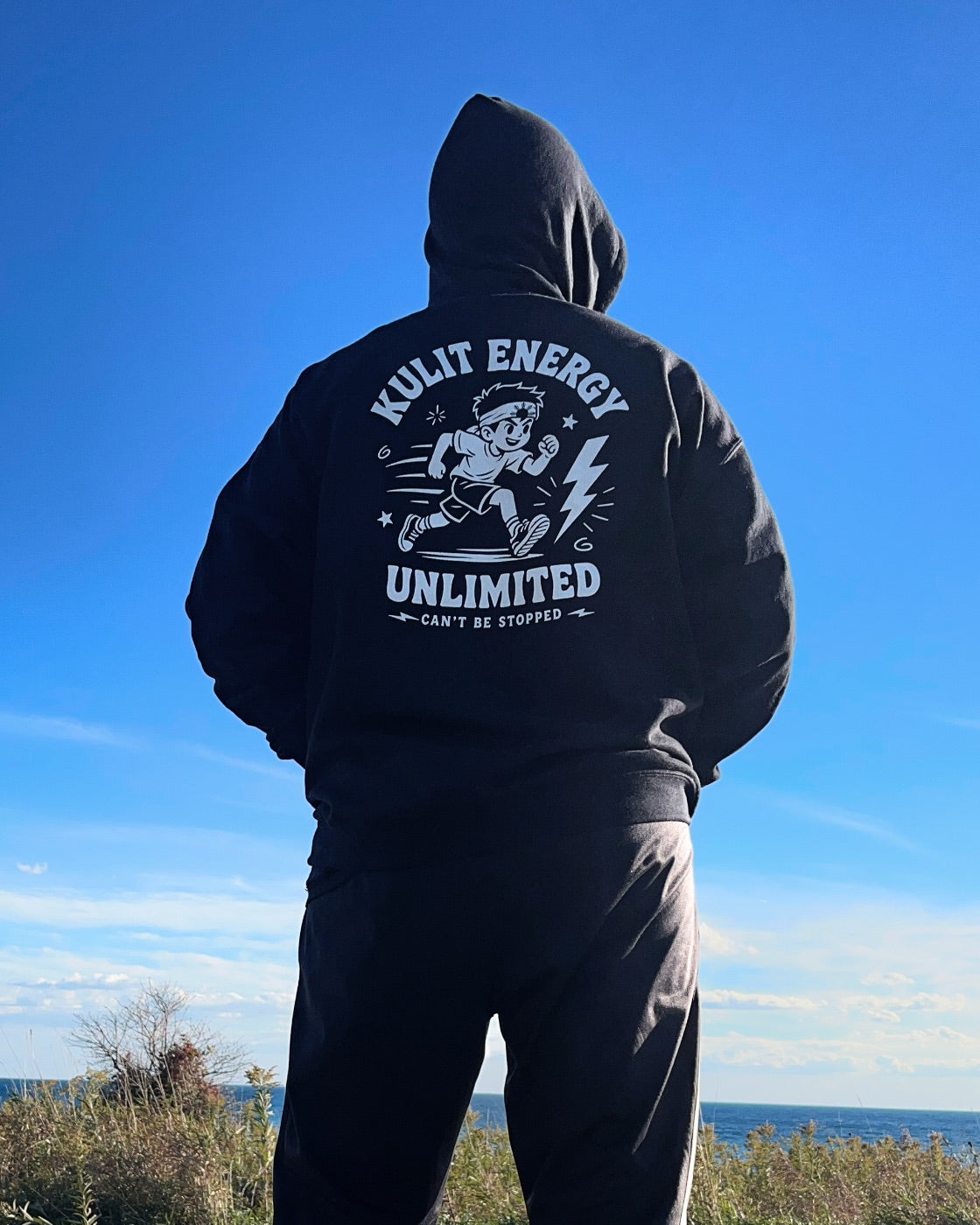 A person viewed from behind wearing a black hoodie and matching black sweatpants, standing outdoors with a large white back graphic that reads 'KULIT ENERGY UNLIMITED' and 'CAN'T BE STOPPED.' The background shows a bright blue sky and a strip of ocean and grass at the horizon.