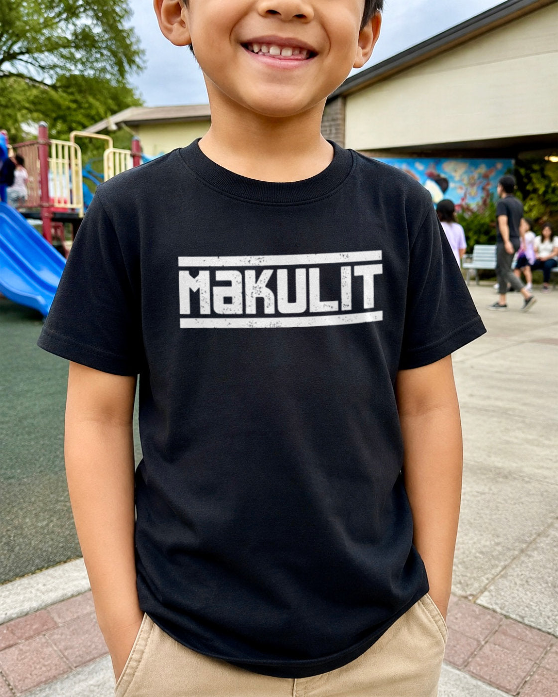 A smiling young boy wearing a black short-sleeve t-shirt with 'MAKULIT' printed in distressed white block letters across the chest, standing outdoors at a playground.