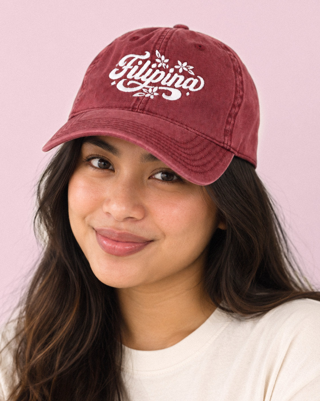 A woman wearing a washed burgundy baseball cap with 'Filipino' embroidered in white script with small flower details, set against a solid light pink background.