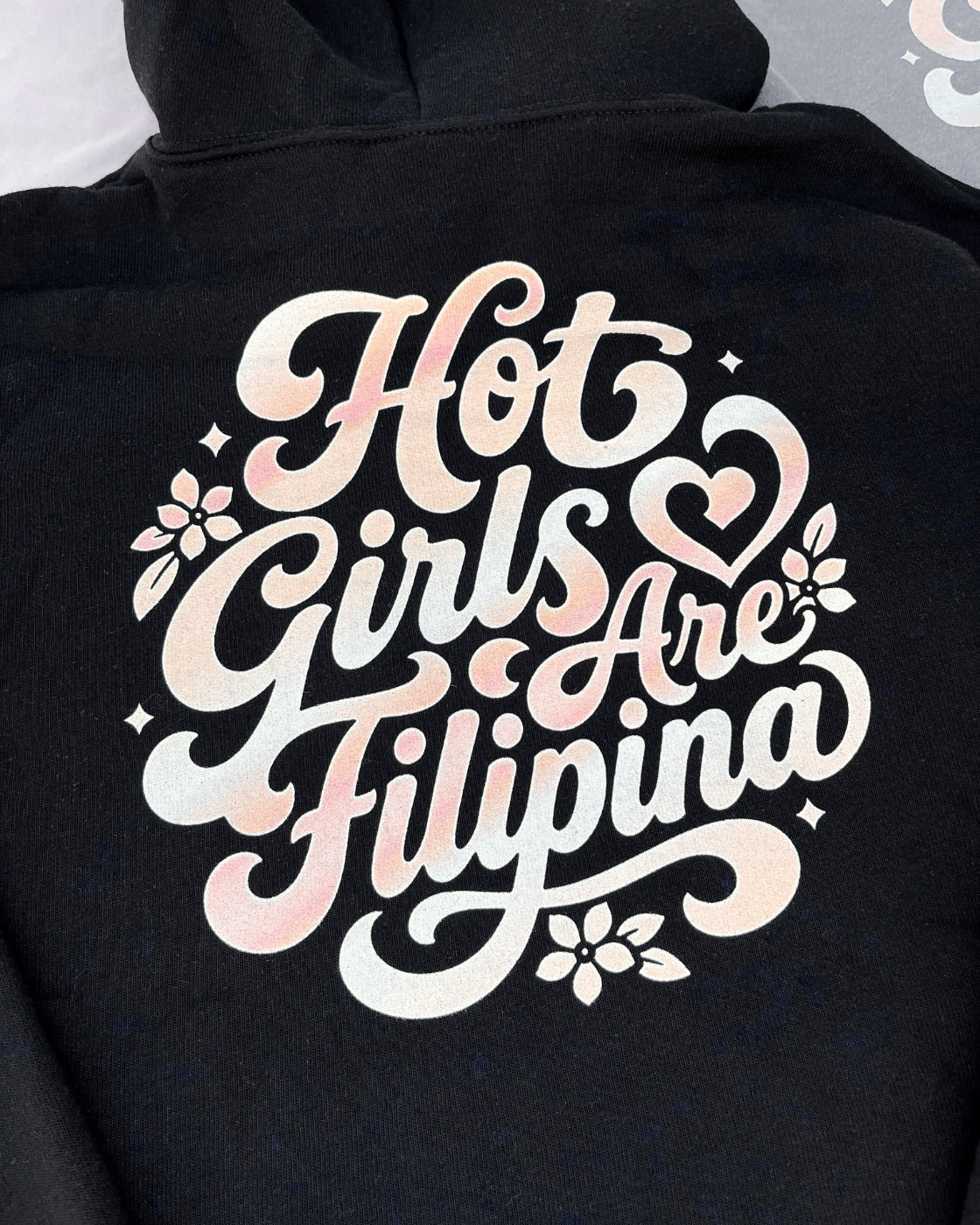 A close-up of a black hoodie featuring a large graphic print on the back with the phrase 'Hot Girls Are Filipina' in a white-to-pale-pink gradient script, decorated with small floral and star details.