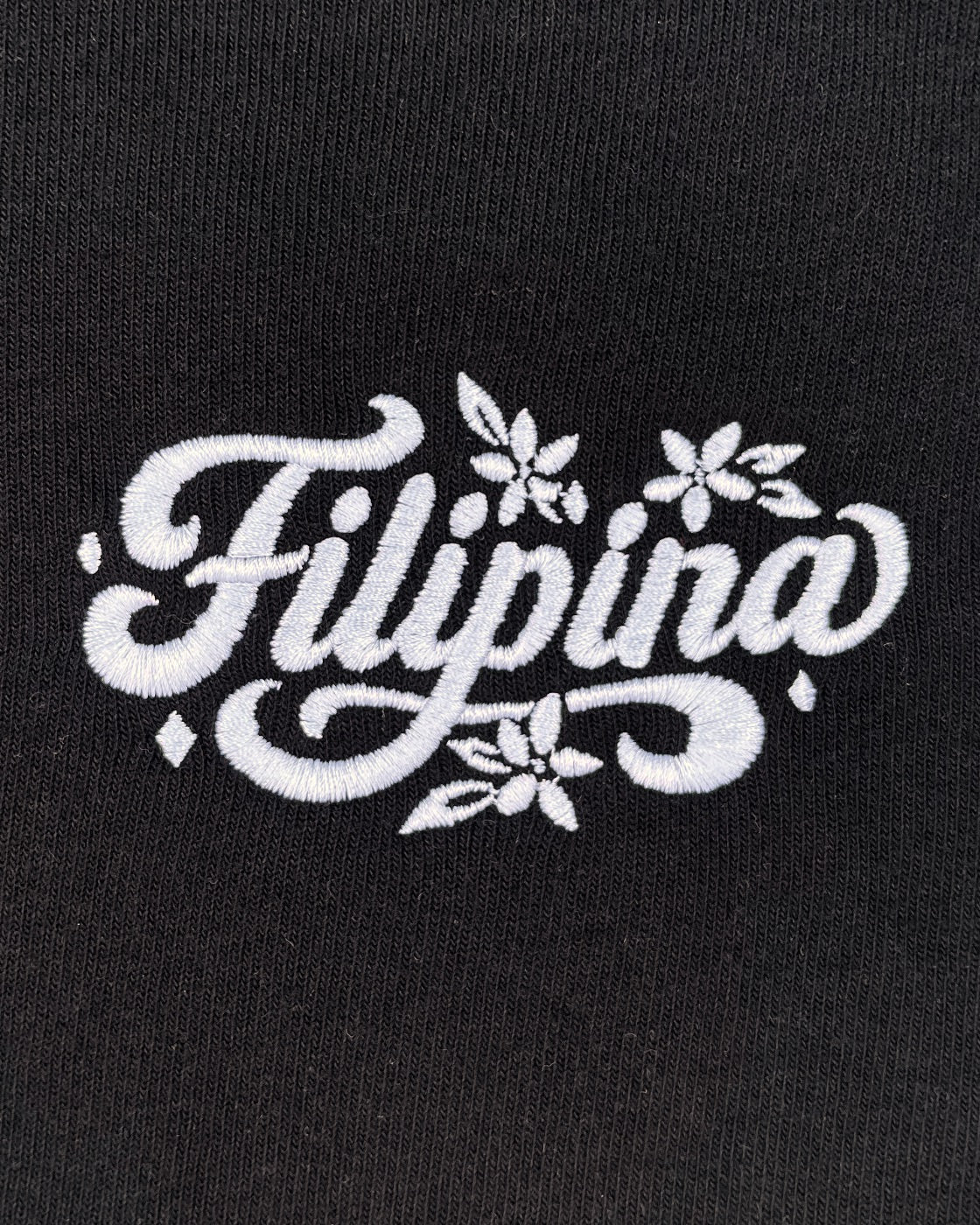 Close-up of a black fabric surface with a white embroidered script reading "Filipina" surrounded by small floral motifs.
