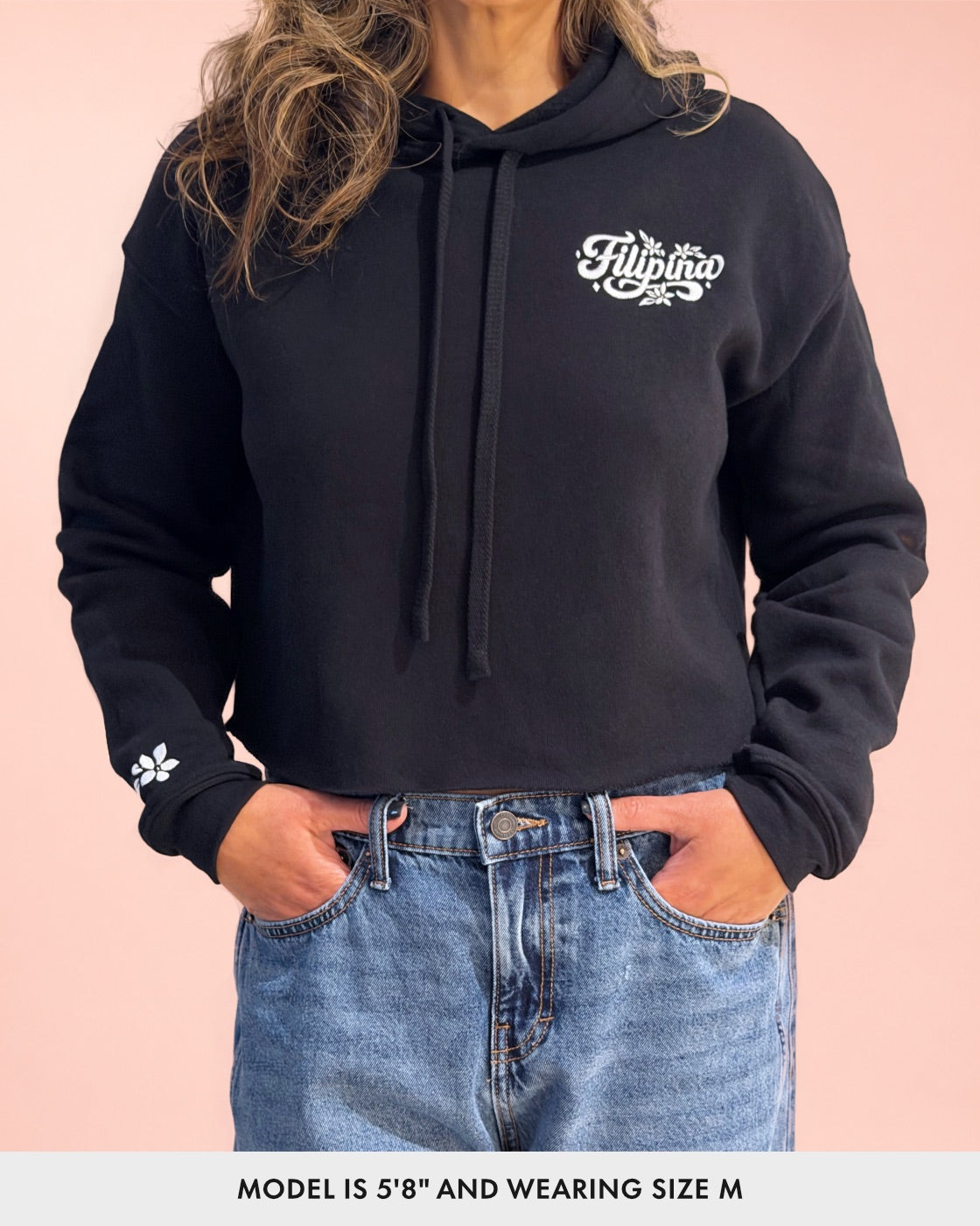 A woman wearing a black cropped hoodie with white 'Filipina' embroidery on the left chest and a small white embroidered sampaguita flower on the right sleeve, paired with blue jeans against a solid pink background.