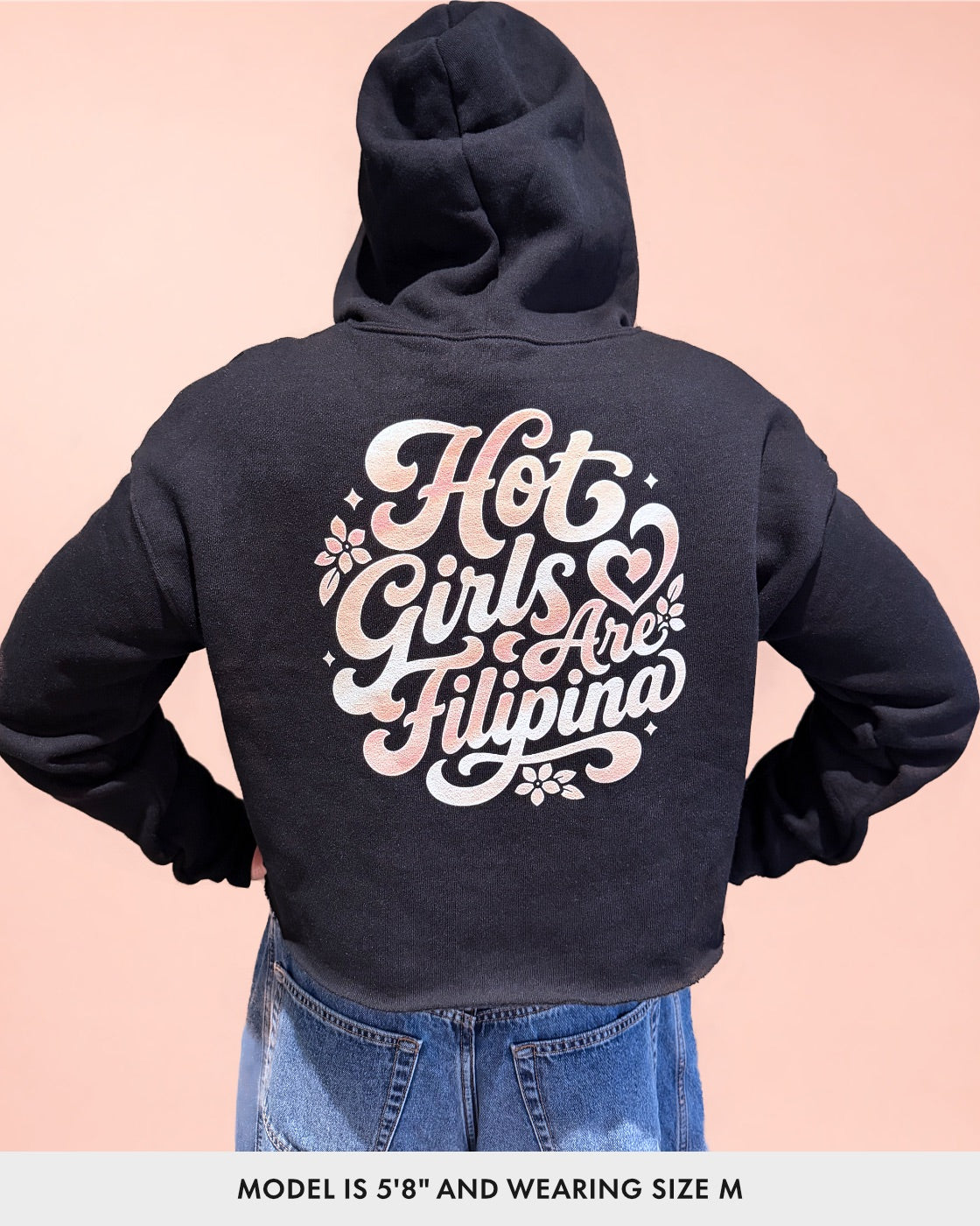 A person shown from the back wearing a cropped black hoodie with a large decorative text graphic on the back reading “Hot Girls Are Filipina” in white and pink lettering, paired with blue jeans, against a pale peach background.
