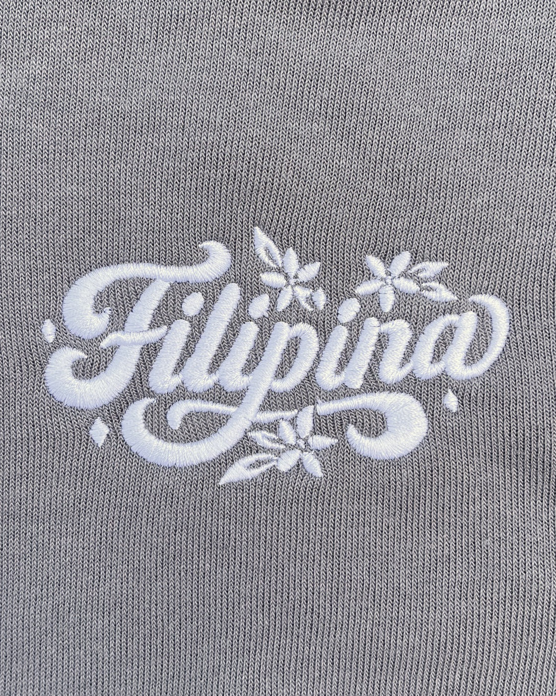 A close-up of a gray fabric featuring white embroidered cursive text that reads 'Filipina' with small floral accents.