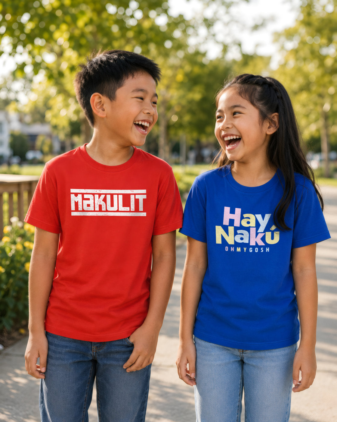 Two children standing outdoors in a park-like setting, each wearing a graphic T-shirt. The child on the left wears a bright red T-shirt with white text reading "MAKULIT," and the child on the right wears a bright blue T-shirt with multicolor text reading "Hay Naku" and smaller text below.