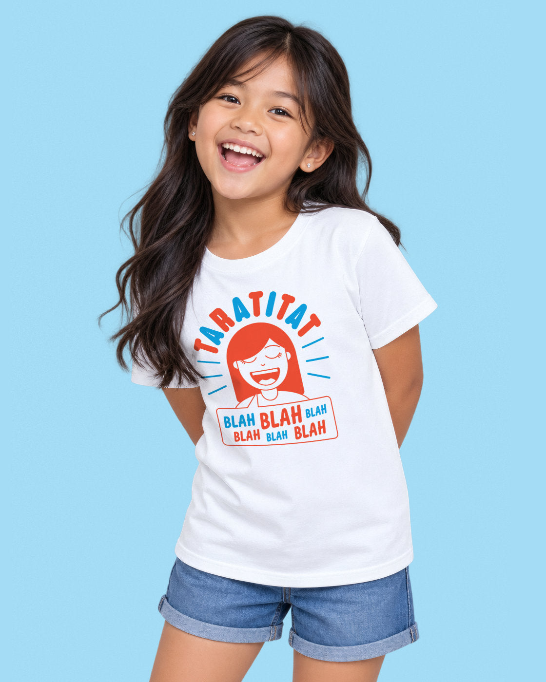 A young Filipino girl with long dark hair is smiling and laughing while wearing a white graphic t-shirt and denim shorts against a solid light blue background. The t-shirt features a colorful cartoon graphic of a girl with red hair and the text 'TARATITAT' and 'BLAH BLAH BLAH'.