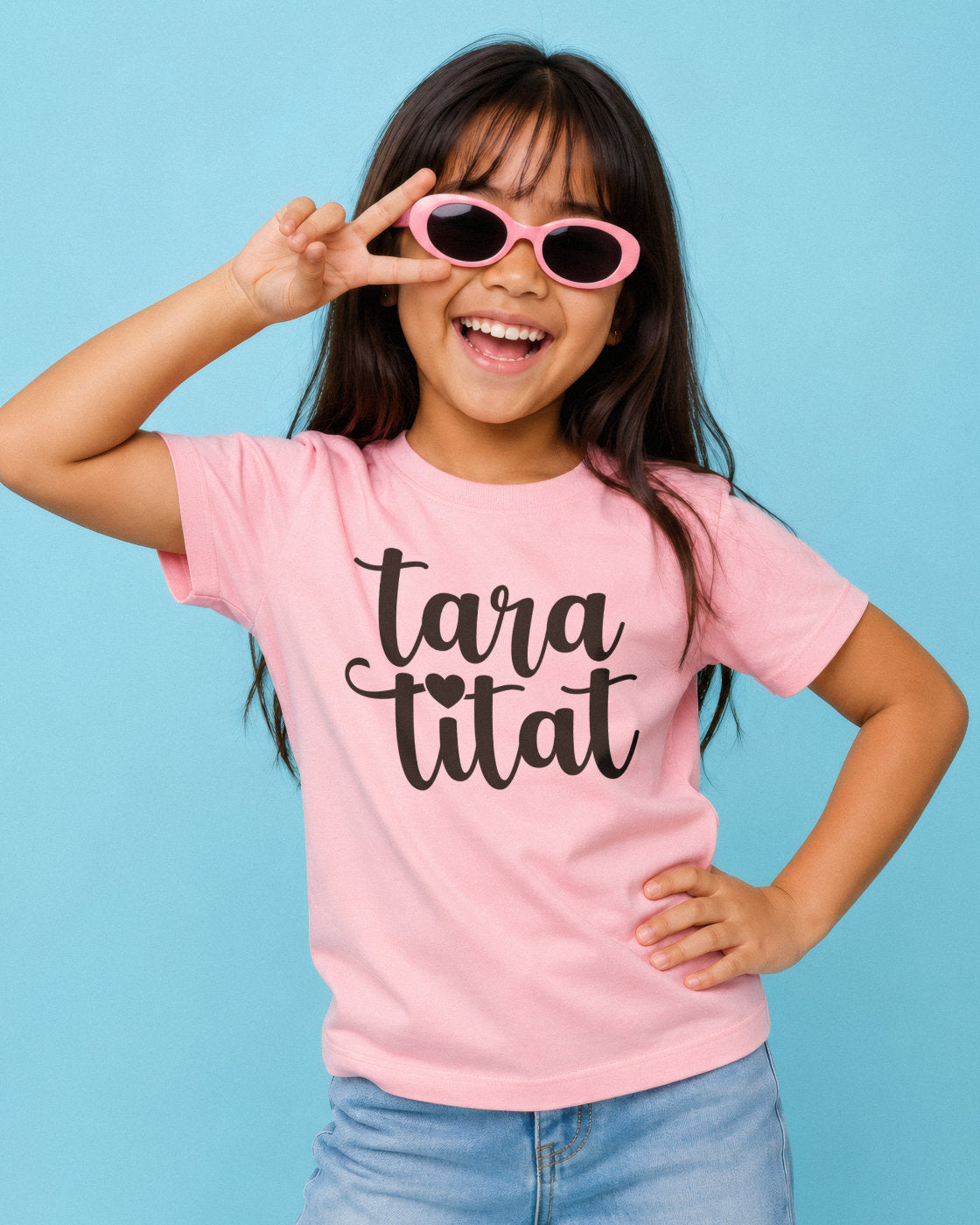 A young Filipino girl with dark hair smiling and posing in a light pink t-shirt with the text 'taratitat' in black script, wearing pink sunglasses, set against a solid light blue background.